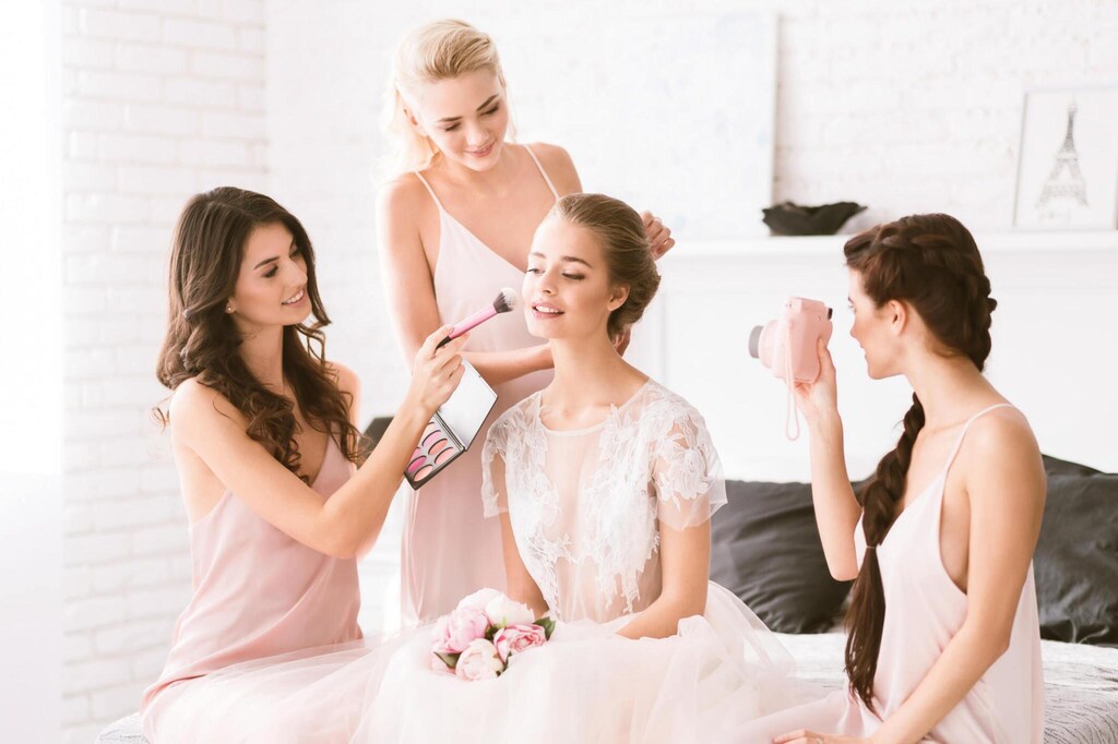 Delighted smiling young bridesmaids sitting in the white bedroom while helping the bride to get ready and expressing joy